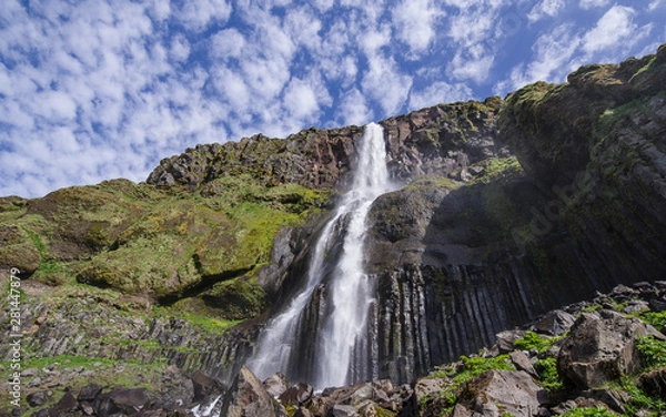 Fototapeta Summer in Iceland - view of a waterfall (Bjarnarfoss) in Snaefellsnes Peninsula. Stream falling from a high black volcanic rock formation. Above blue sky dotted by white clouds. Long exposure . 