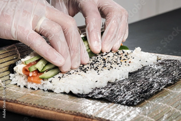 Fototapeta Cook's hands close-up. A male chef makes sushi and rolls from rice, red fish and avocado. White gloves.