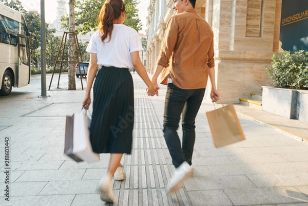 Fototapeta Lower body section of a young tourist couple walking by store windows and holding paper shopping bags in a destination city.