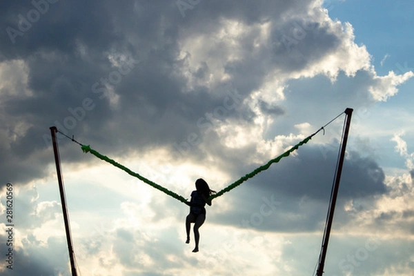 Fototapeta Girl jumping on an extreme swing. The girl flies launched by a rubber slingshot. The girl is experiencing adrenaline and joy from the jump. Woman on a background of clouds in flight.