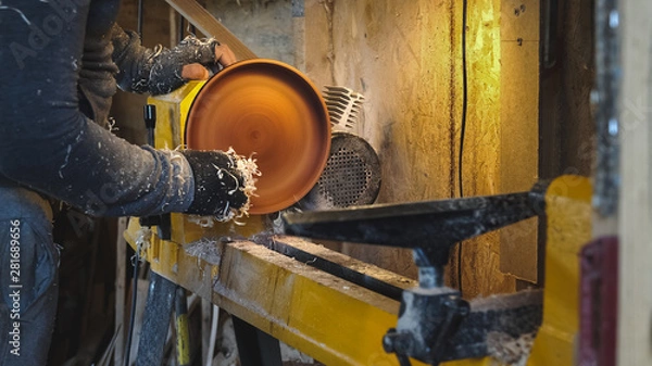 Fototapeta Woodworker grinds the workpiece by sawdust. Carpenter completes work at lathe. Making beautiful wooden dishes handmade. Builder works with workpiece in workshop. Creation unusual dishes.