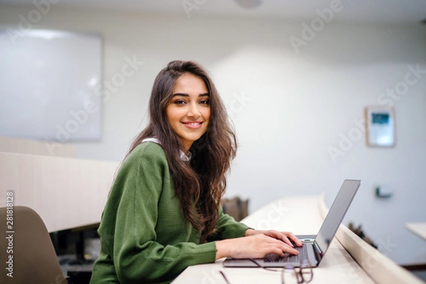 Obraz Portrait of a beautiful, young and intelligent-looking Indian Asian woman student wearing a white shirt and green tracker smiling as she works on her laptop in a university classroom.