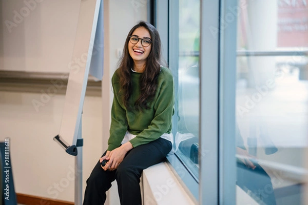 Fototapeta Portrait of a young, educated, confident and intelligent-looking Indian Asian student girl smiling as she sits in a seminar room by the window during the day in a preppy outfit.