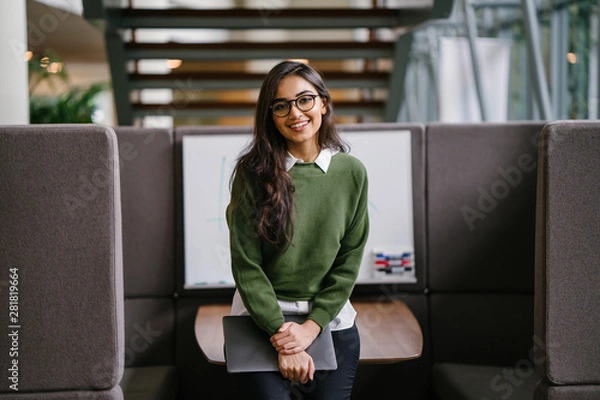 Fototapeta Portrait of a young, beautiful, intelligent and attractive Indian Asian MBA student smiling as she leans in a discussion booth in her campus. She is wearing a preppy outfit and smiling confidently.