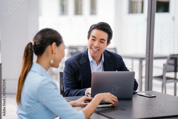 Fototapeta A young, handsome and well-groomed Asian man in a dark suit is interviewing a professional woman candidate applying a job during the day. He is smiling as he talks to the woman.