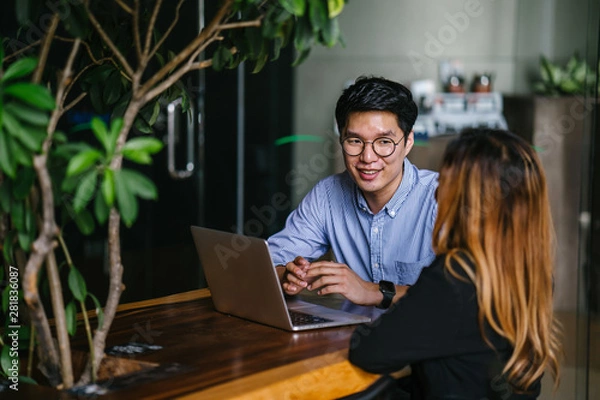 Fototapeta A pair of diverse team mates have a business meeting and discussion together to collaborate. A Korean man is sitting at a table in a trendy coworking space office and talking to his female companion.