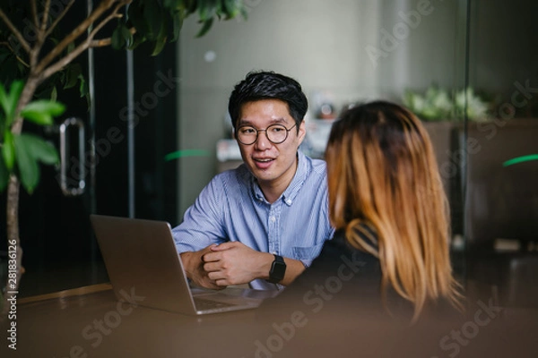 Fototapeta A pair of diverse team mates have a business meeting and discussion together to collaborate. A Korean man is sitting at a table in a trendy coworking space office and talking to his female companion.