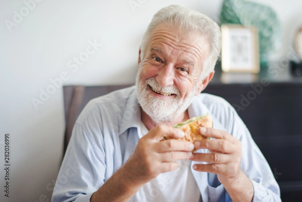 Fototapeta Seniors European man is sitting to eat a burger at home which looking at camera. Retired man is smiling with felling happy and take a burger on hand.