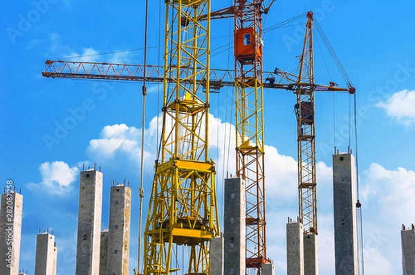 Fototapeta Yellow and Red Cranes, Vertical Reinforced Concrete Columns with Rebars against Blue Sky and White Clouds. Real Estate, Residential Buildings Urban Mixed-Use Development Concept.