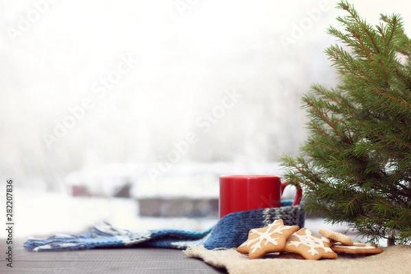 Fototapeta holiday cookies and a mug with a scarf under the tree against the background of a landscape with snowy houses. cozy winter morning