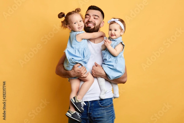 Fototapeta happy awesome daddy holding his favourite daughters in hands, enjoying time with kids, family traditions. Love, deep affection and devotion. isolated yellow background