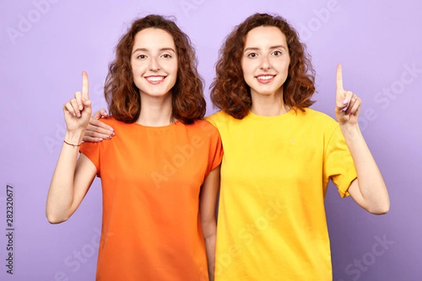 Fototapeta Happy beautiful twins girls point up isolated on blue background, two sisters showing something above their heads , advertisement, place for text, body language