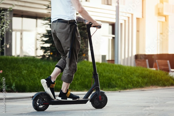 Fototapeta Close up of man riding black electric kick scooter at beautiful park landscape