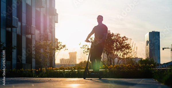 Obraz Man riding black electric kick scooter at cityscape background at sunset