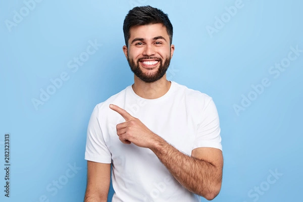 Fototapeta Young handsome charming man showing something with index finger, place for text, advert, copy space. isolated on blue background. direction. body language. sale, shop