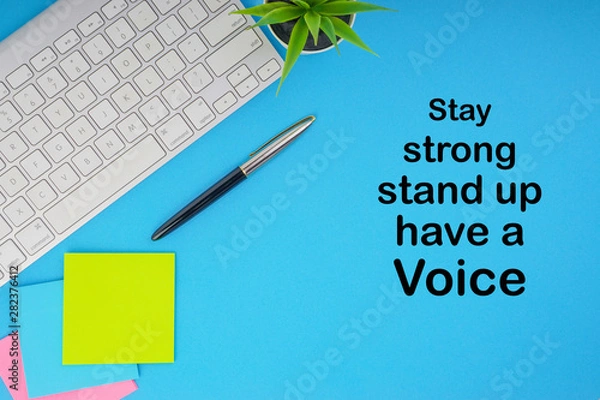 Obraz Flat lay, top view office table desk. Workspace with STAY STRONG STAND UP HAVE A VOICE inscription written with keyboard, pen and sticky notes on blue background. Business, Insurance and motivation co