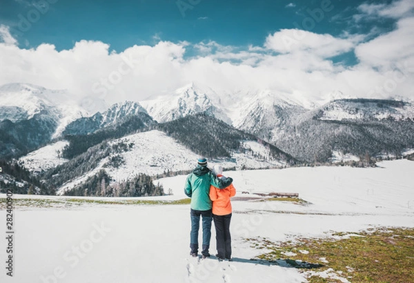 Obraz Loving couple standing and looking on beautiful snowy mountains peaks. Panoramic view. Back view.