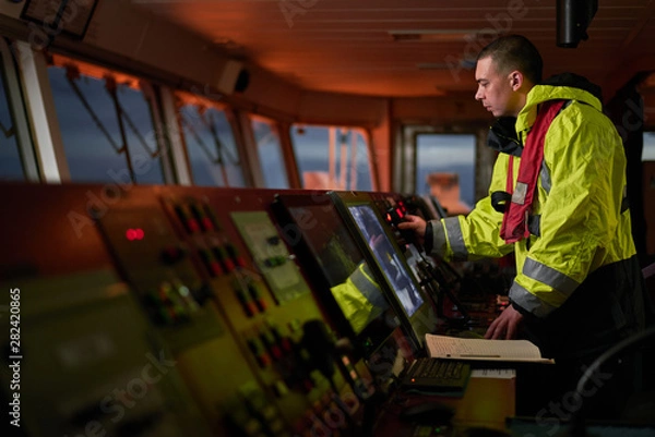 Obraz Navigator. pilot, captain as pat of ship crew performing daily duties with VHF radio, binoculars on board of modern ship with high quality navigation equipment on the bridge on sunrise.