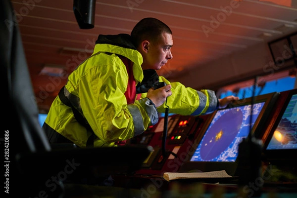 Obraz Navigator. pilot, captain as pat of ship crew performing daily duties with VHF radio, binoculars on board of modern ship with high quality navigation equipment on the bridge on sunrise.