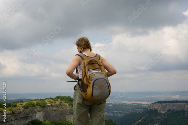 Obraz a girl-traveler in a backpack looks into the distance from a cliff on the ruins of a medieval fortified city