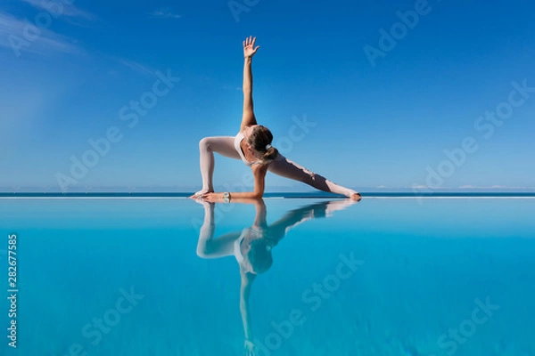 Obraz Charming woman yoga lover in overalls makes Uthit Triconasana by the pool while relaxing at sea in a sunny warm country. The concept of relaxation and better health. Copyspace