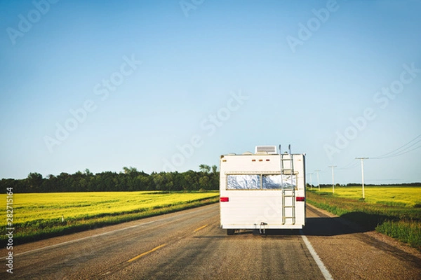 Fototapeta The backside of an older pull type camper moving down a highway between canola fields in a countryside summer landscape