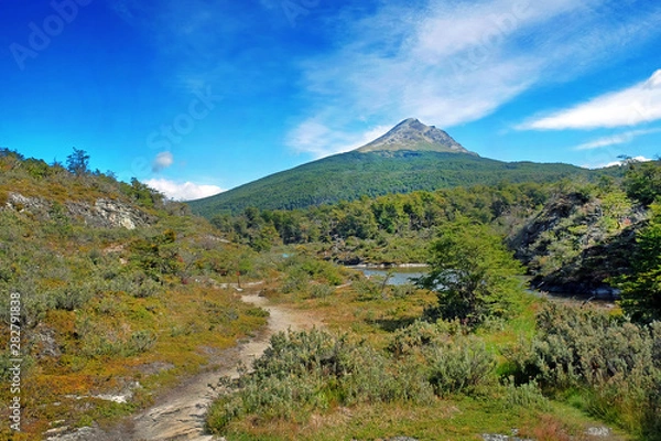 Fototapeta Panoramic view of Tierra del Fuego National Park, showing hills and a volcano surrounded by green vegetation and water, against a blue sky.