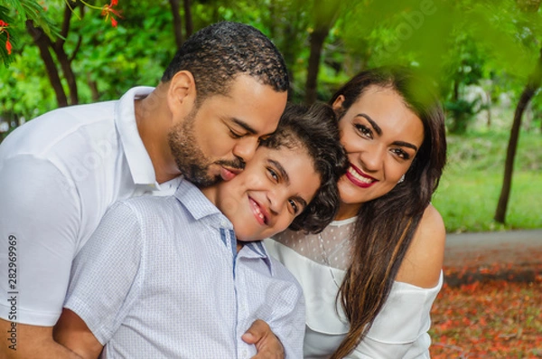 Obraz couple of Latin American men and women, with boy suffers autism, happy in a portrait family outdoors together in a park, the three laughing hugging