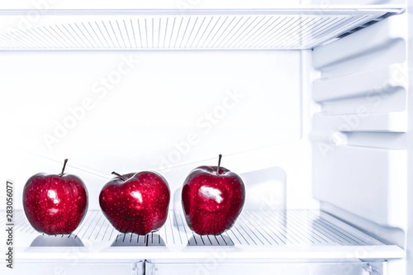 Fototapeta Three red apples is lying on the empty shelf of the fridge. Diet and weight loss concept.