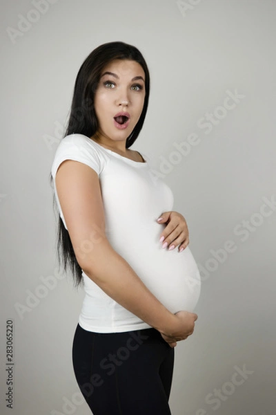 Fototapeta beautiful pregnant brunette woman holding her pregnant belly with surprise on isolated white background