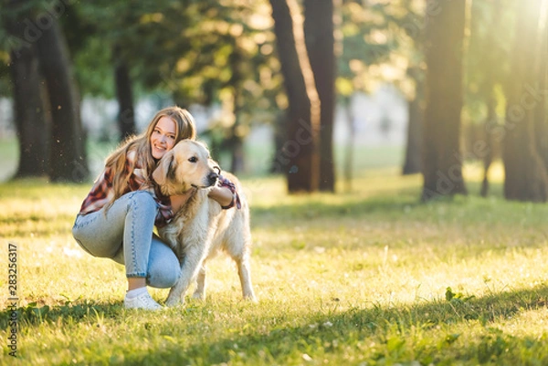 Fototapeta full length view of beautiful young girl in casual clothes hugging golden retriever while sitting on meadow in sunlight and looking at camera