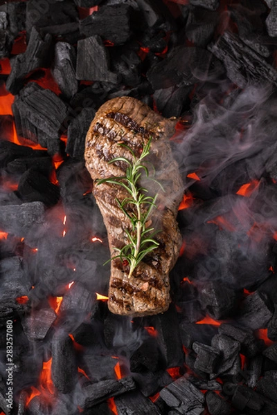 Fototapeta Fried meat steak on a black background of charcoal. Cooked juicy steak with smoke on the coals with rosemary. Grilled delicious marinated steak on coals, closeup photo. Top view