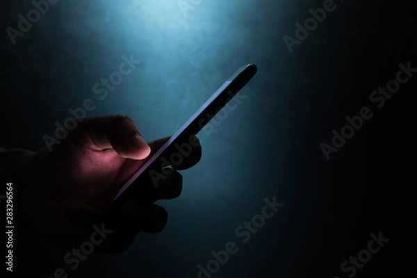 Obraz silhouette hand holding and touching a mobile phone screen in the dark darkness against dark blue background