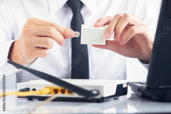 Fototapeta Male IT support holding RJ11 cable connecting ADSL Splitter with modem and laptop on white table