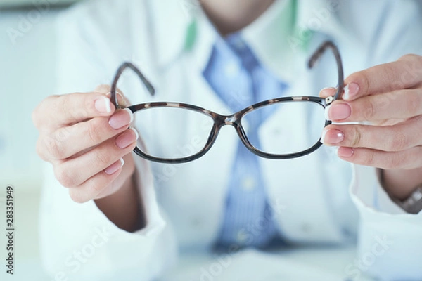 Fototapeta Female optician hands giving new glasses to customer for testing and trying close-up. Eye doctor with client comparing spectacles and choosing lenses in store.