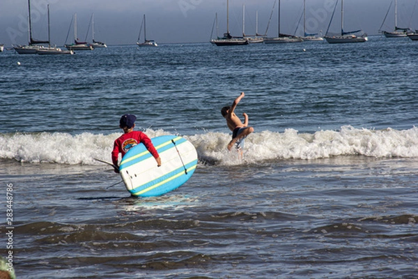 Fototapeta Swimmers heading into the ocean, 1 of them carrying a surfboard. White cap waves, and sailboats floating on the horizon.