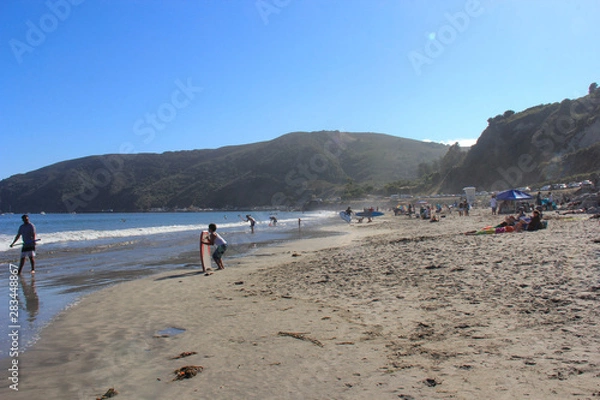Fototapeta Crowds playiing on the beach. Mountains in the background with clear blue skies.