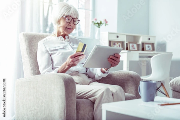 Fototapeta Modern retired woman holding bank card while shopping online via tablet