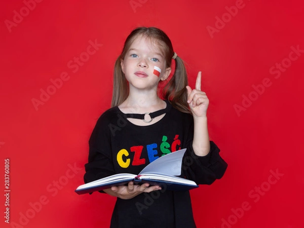 Fototapeta little girl in a black sweatshirt, on her sweatshirt the word Hello in Polish raised her finger up, holds a notebook or book in her hand, Advertising concept for learning Polish