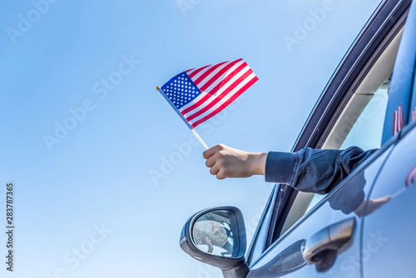 Fototapeta Chłopiec trzyma flagę Ameryki z otwartego okna samochodu na tle nieba. USA. Koncepcja