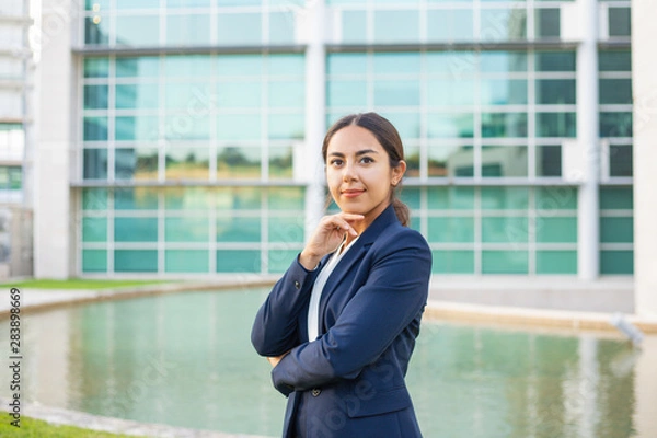 Fototapeta Satisfied successful business leader posing outside. Beautiful Latin woman wearing formal suit, standing near office building, touching chin and looking at camera. Young business woman concept