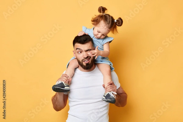 Fototapeta niegrzeczne zachowanie. rodzic nie radzi sobie z dzieckiem z dużą energią. dziewczyna dotyka oczu taty, siedząc na ramionach. zbliżenie zdjęcia. pojedyncze żółte tło. niedopuszczalne zachowanie
