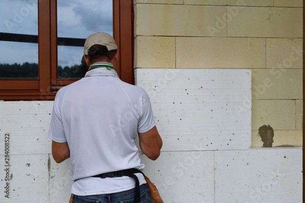 Obraz Worker installing white rigid polyurethane foam sheet on aerated concrete block building wall facade for energy saving. Diy, house improvement and insulation concept.