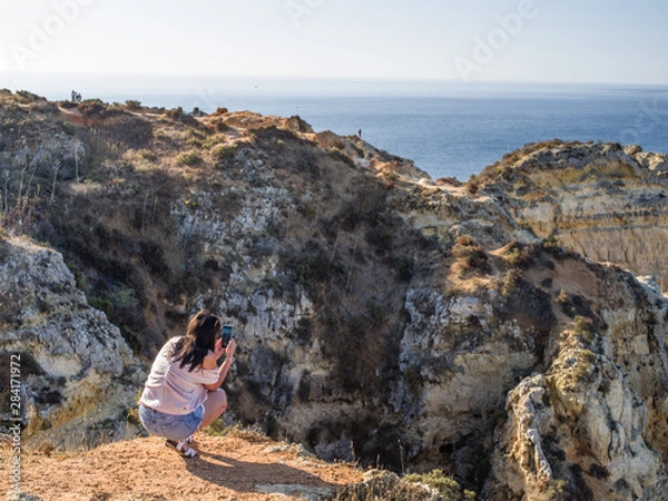 Obraz Young lady taking a photo of the colourful cliffs of Ponta da Piedade in Algarve. Mornuing (low) sun illuminated, lady and cliffs in foreground, ocean and sky in background