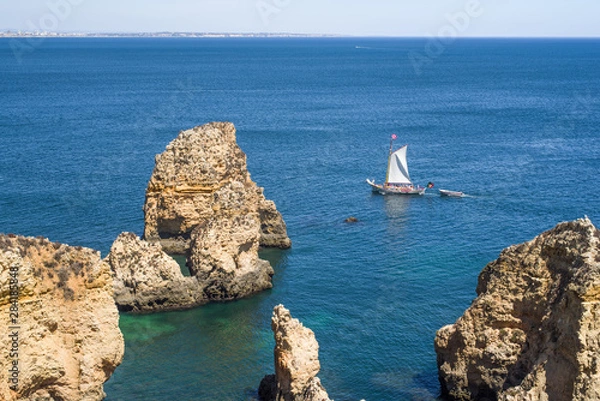 Fototapeta Duża żaglówka z małą łodzią przechodzącą obok kolorowych klifów Ponta da Piedade w Algarve. Zdjęcie zrobione z góry. Skały i turkusowe woda morska na pierwszym planie, miasto Lagos na odległość