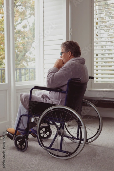 Fototapeta Disabled active senior man looking through window on wheelchair in a comfortable home