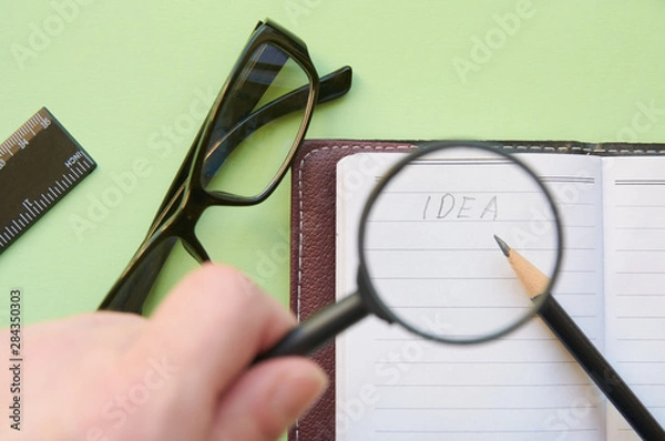 Fototapeta The inscription "Idea" under the magnifying glass in the hands of a person. Idea search concept. Flat lay