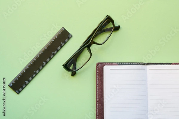 Fototapeta Conceptual layout on a green background. A detailed notebook, glasses and a ruler. Flat lay.