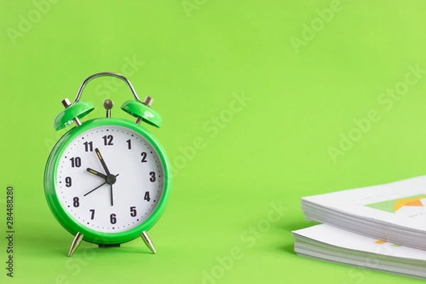 Fototapeta Green alarm clock with stack of financial documents on green background