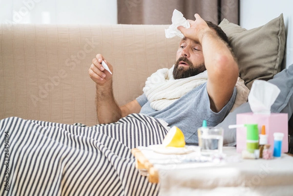 Fototapeta High temperature concept. Man feels badly ill. How to bring fever down. Cold symptoms and causes. Sick man with flu. Man hold thermometer. Measure temperature. Break fever remedies.